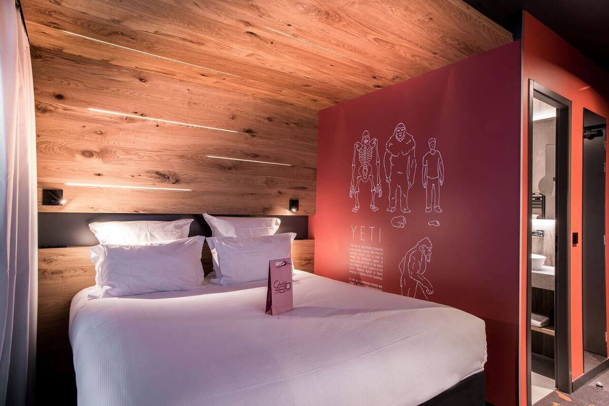 A modern hotel room with a wooden ceiling, a large bed with white bedding, a red accent wall with a skeleton illustration, and a small bedside table with a pink item.
