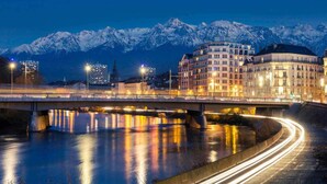 Un puente sobre un río con reflejos de luces, una cordillera nevada al fondo y un edificio de varios pisos con ventanas iluminadas.
