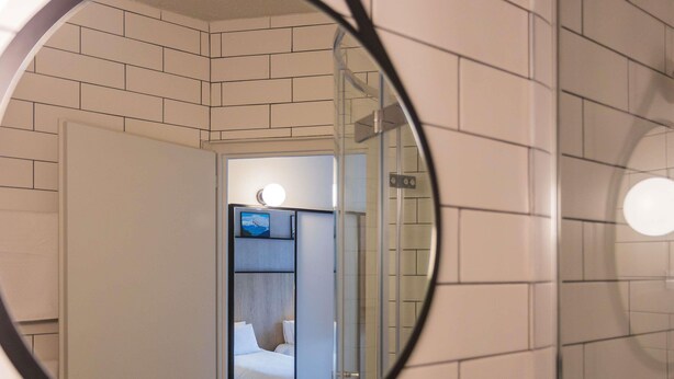 Un baño con un espejo que refleja una habitación con una cama y una mesita de noche.