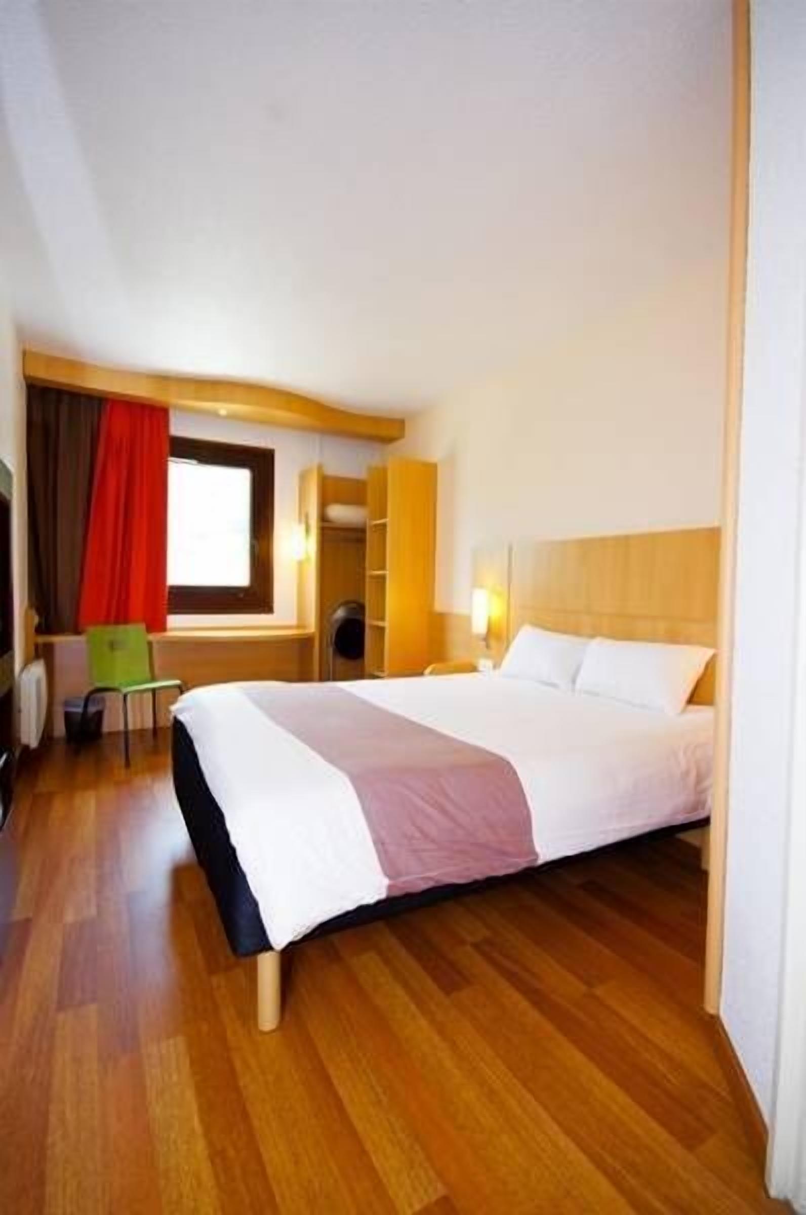 Una habitación de hotel con cama, suelo de madera, ventana con cortinas, escritorio y silla.