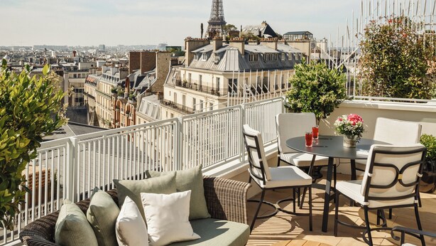 Une terrasse sur le toit, aménagée avec un canapé, des fauteuils et une table, d’où l’on peut admirer une vue panoramique sur la ville, avec une tour imposante en évidence.
