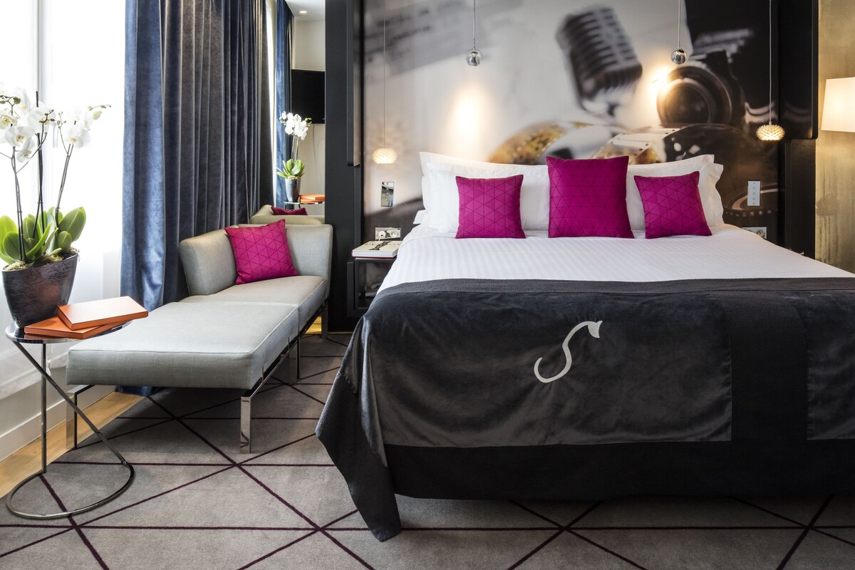 A modern hotel room with a large bed, a sofa, a small table, and a potted plant.