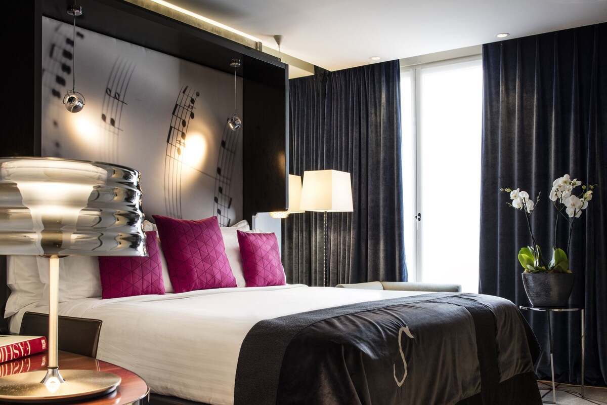 A modern hotel room with a large bed, a bedside table, a lamp, and a potted plant.