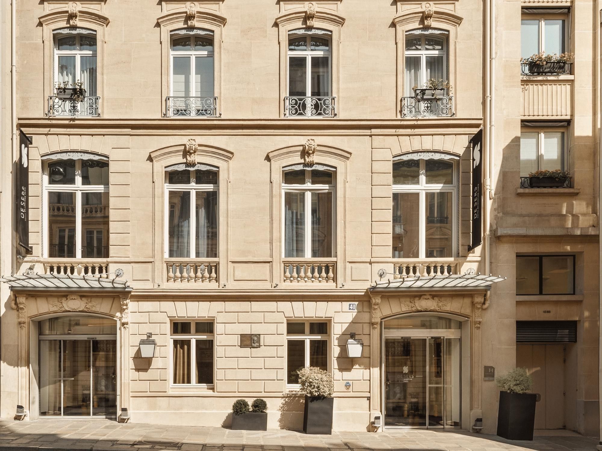 Un edificio de varios pisos con grandes ventanales, balcones decorativos y una entrada principal a nivel de calle con puerta y una pequeña jardinera.
