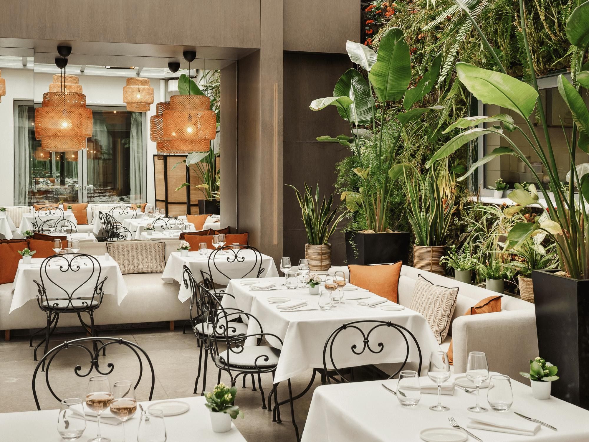Ein Restaurant mit weißen Tischtüchern, schwarzen Metallstühlen und hängenden Rattanlampen.