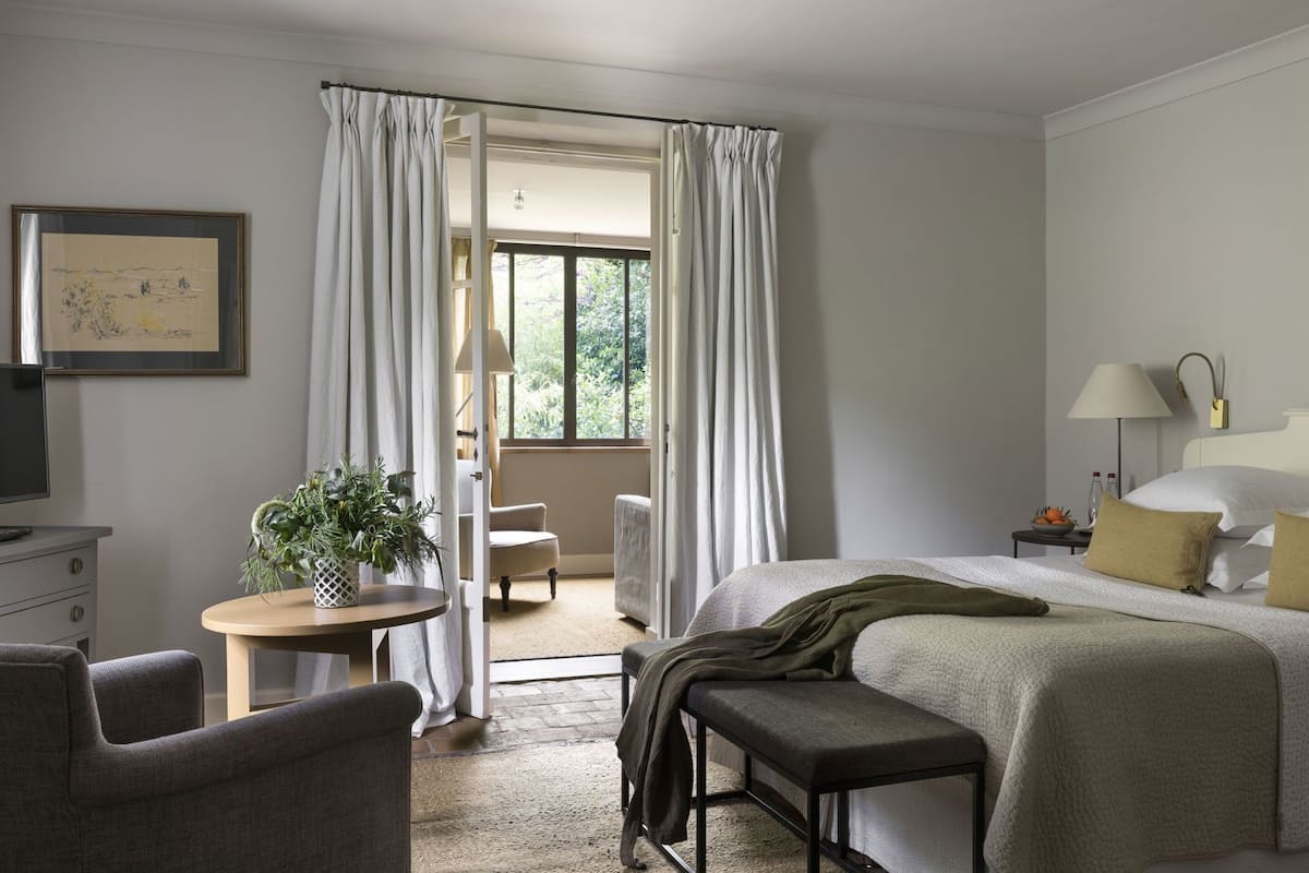 A hotel room with a bed, a desk, a chair, a small table with a plant, and a window with curtains.