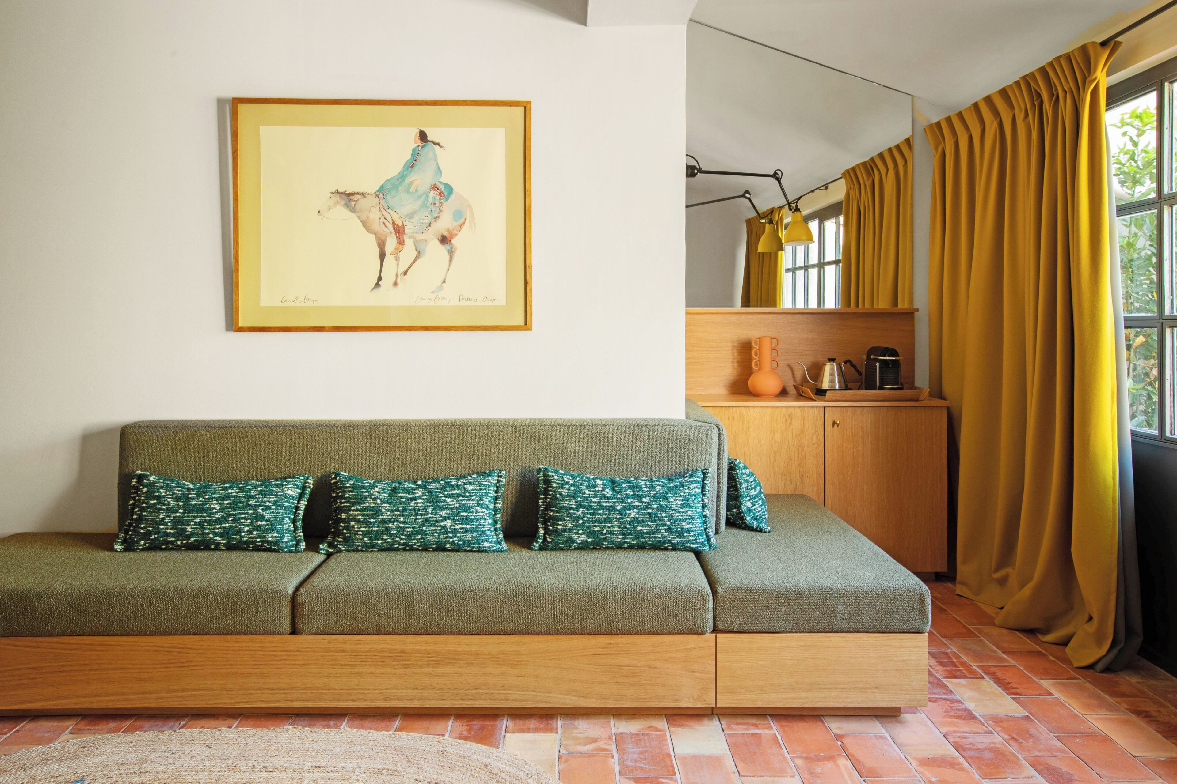A room with a green sofa, a framed painting, a wooden cabinet, and a window with yellow curtains.
