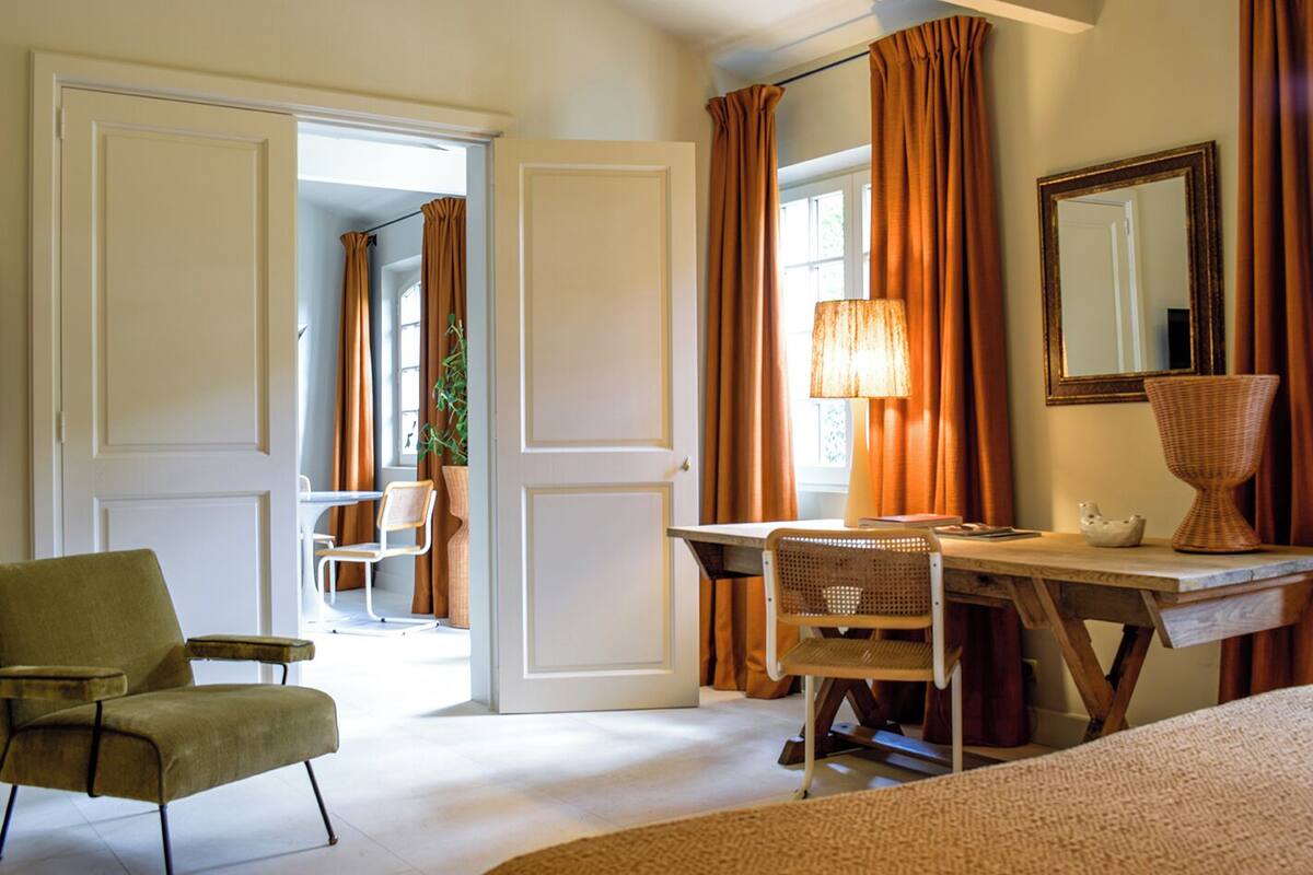A hotel room with a desk, two chairs, a lamp, a mirror, and a bed.