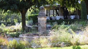 Un jardín con una fuente de piedra, abundante vegetación y un pabellón al fondo.