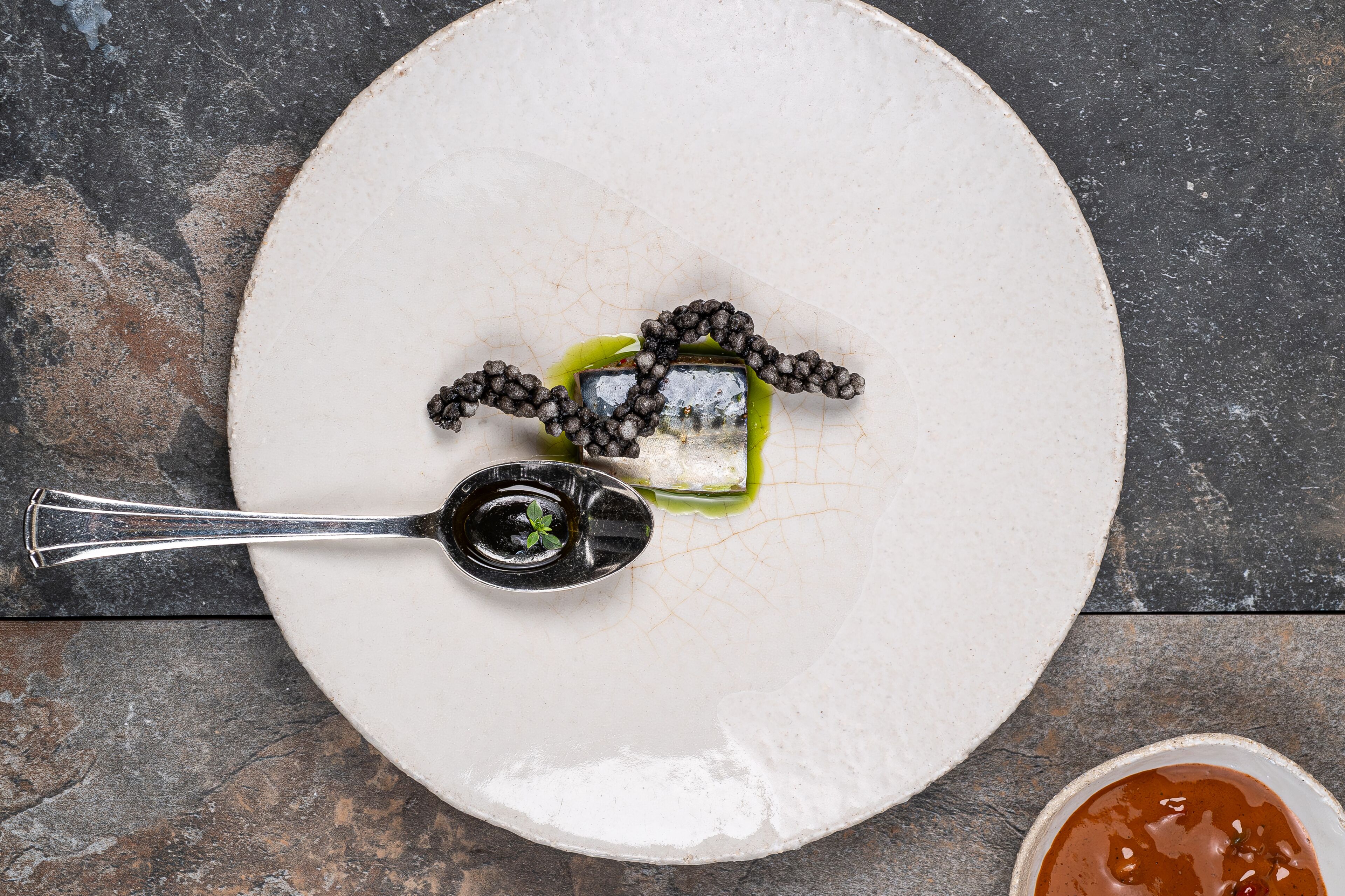 Une assiette blanche avec un petit ramequin de sauce, une cuillère et un morceau de nourriture, le tout posé sur une surface sombre.