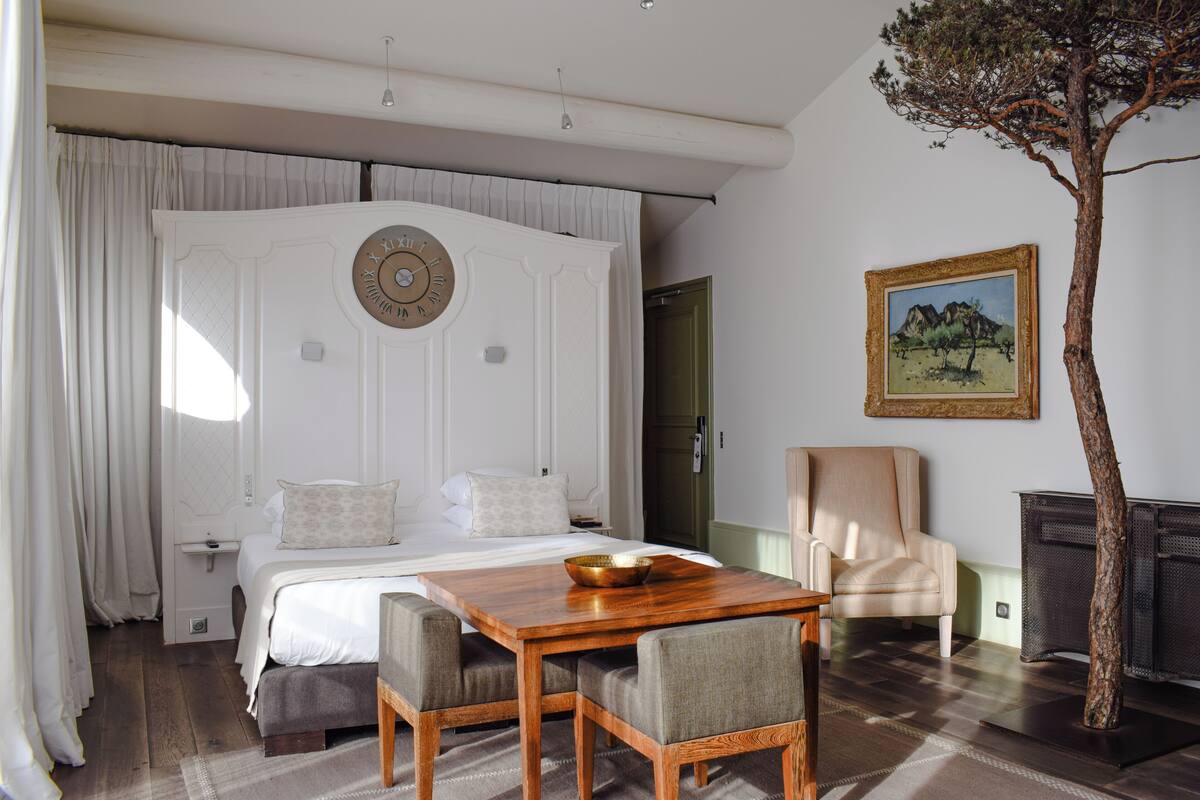 A bedroom with a large bed, a wooden table, a chair, a painting, and a tree sculpture.