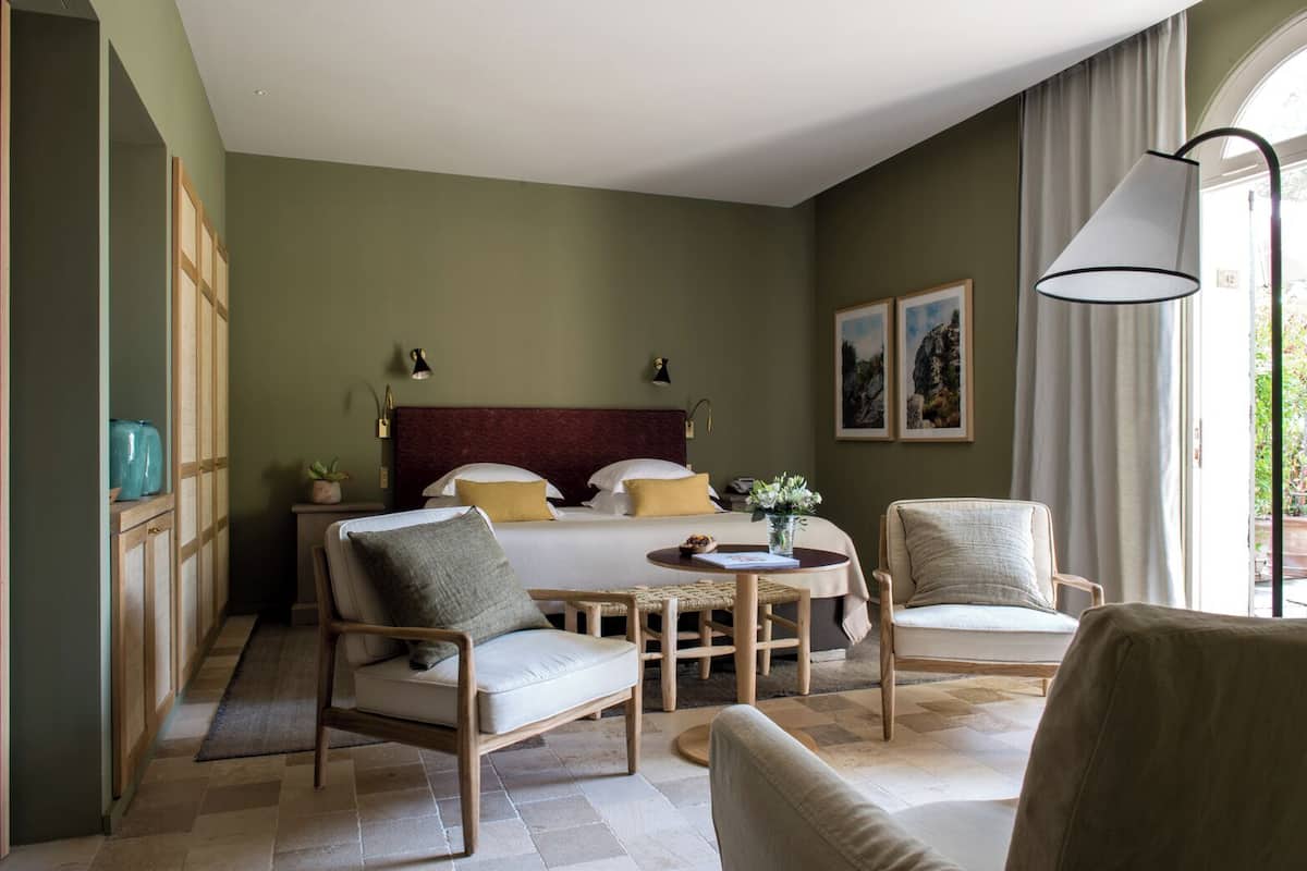 A hotel room with a bed, two chairs, a small table, a lamp, and a view of the outdoors.