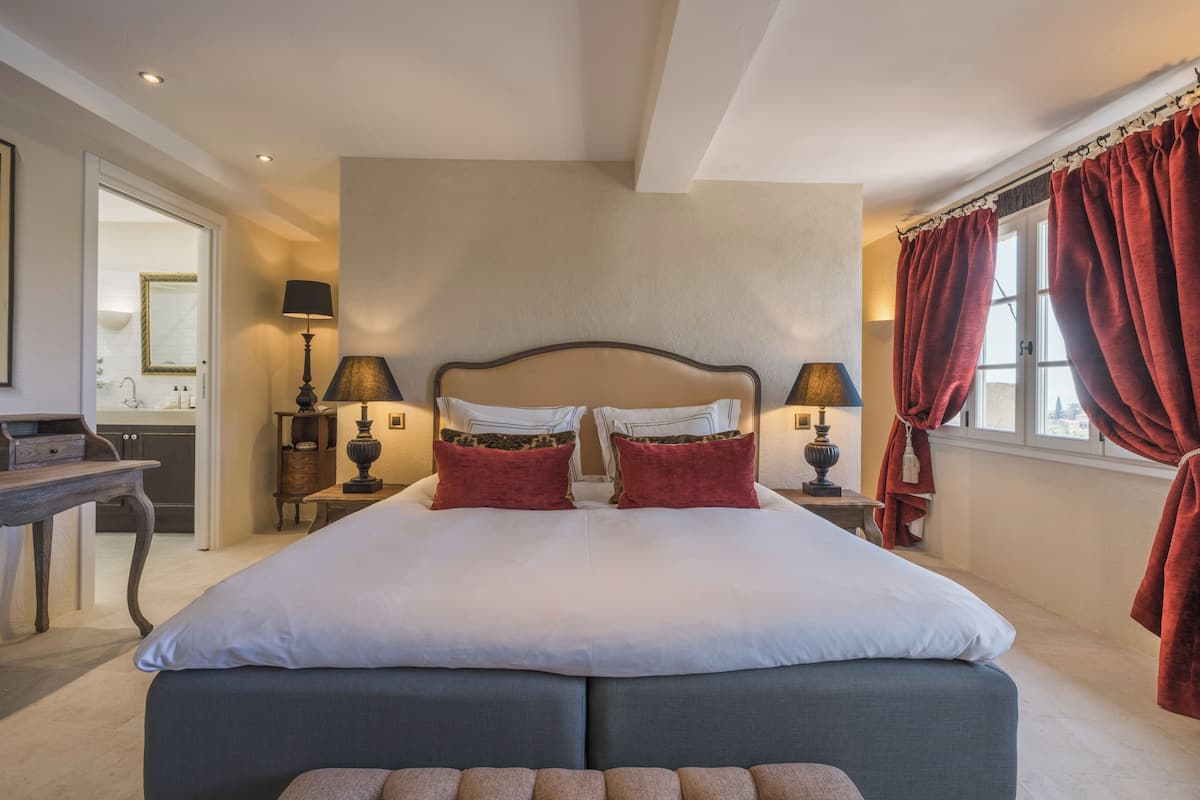 A bedroom with a large bed, two bedside tables with lamps, a wooden nightstand, and a view of the outside through a window with red curtains.