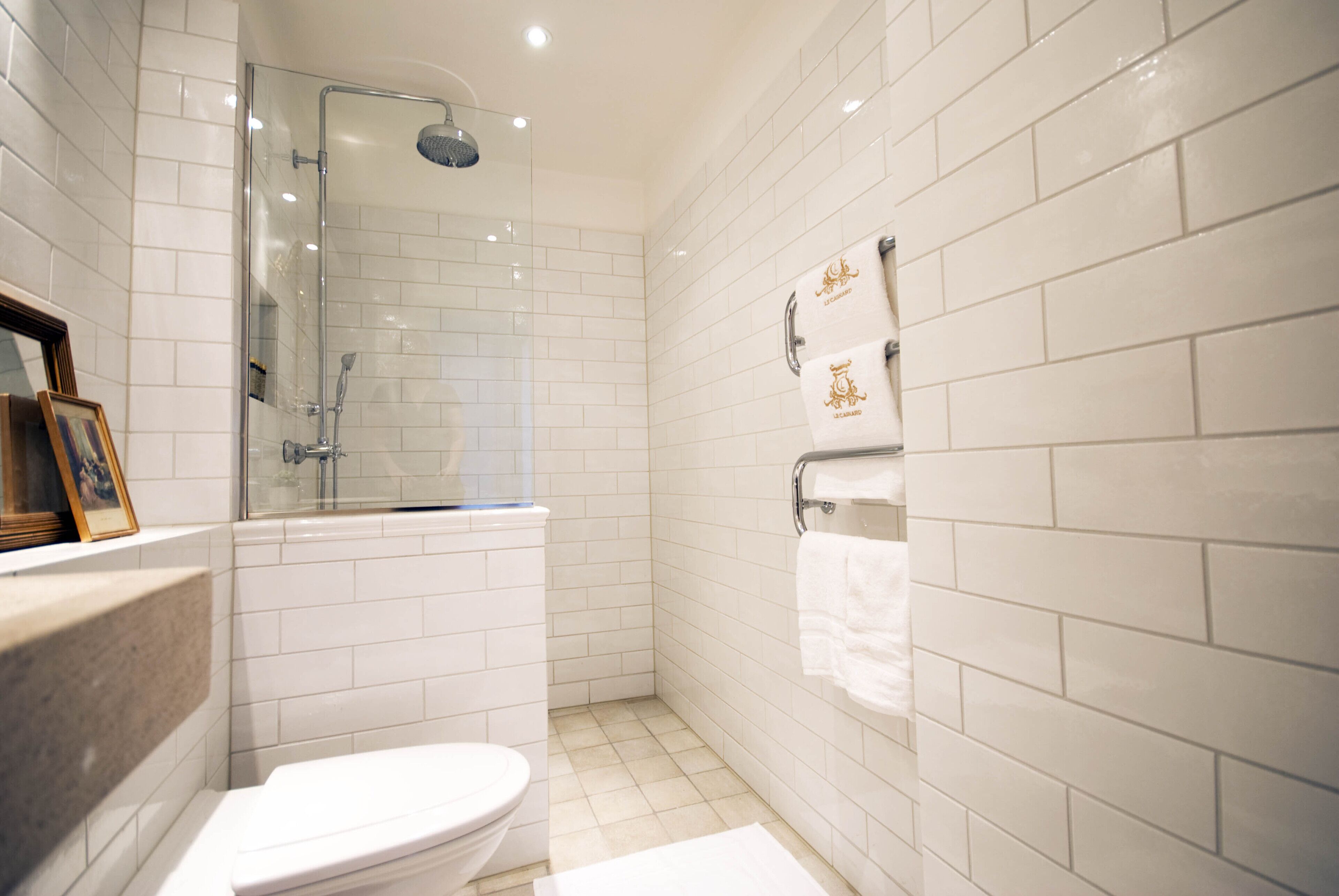 A bathroom with white tiled walls, a glass shower enclosure, a toilet, and a towel rack with two towels.