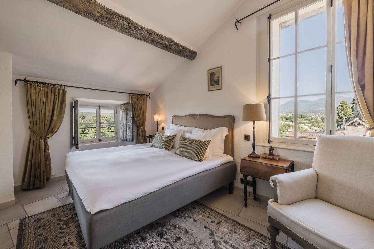A bedroom with a large bed, a nightstand, a chair, and a window with a view of greenery.