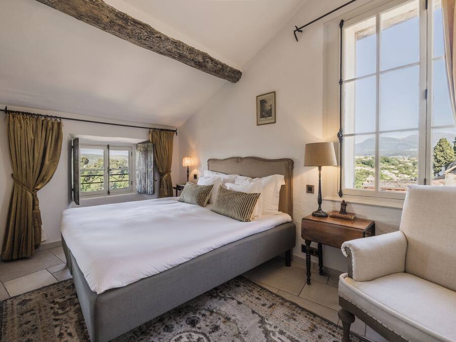 Une chambre à coucher avec un grand lit, une table de chevet, une chaise et une fenêtre donnant sur un paysage verdoyant.