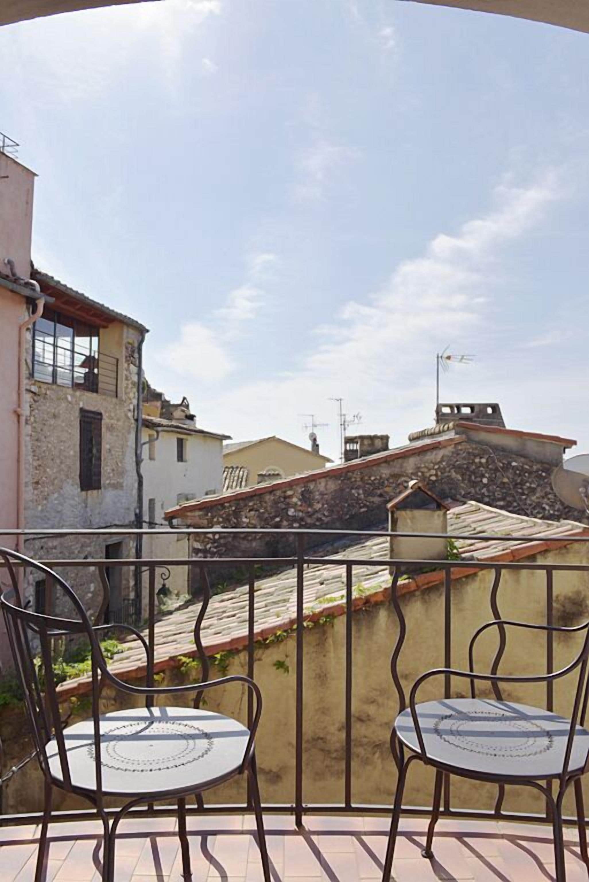 A balcony with two metal chairs and a table, overlooking a residential area with stone and tiled roofs.