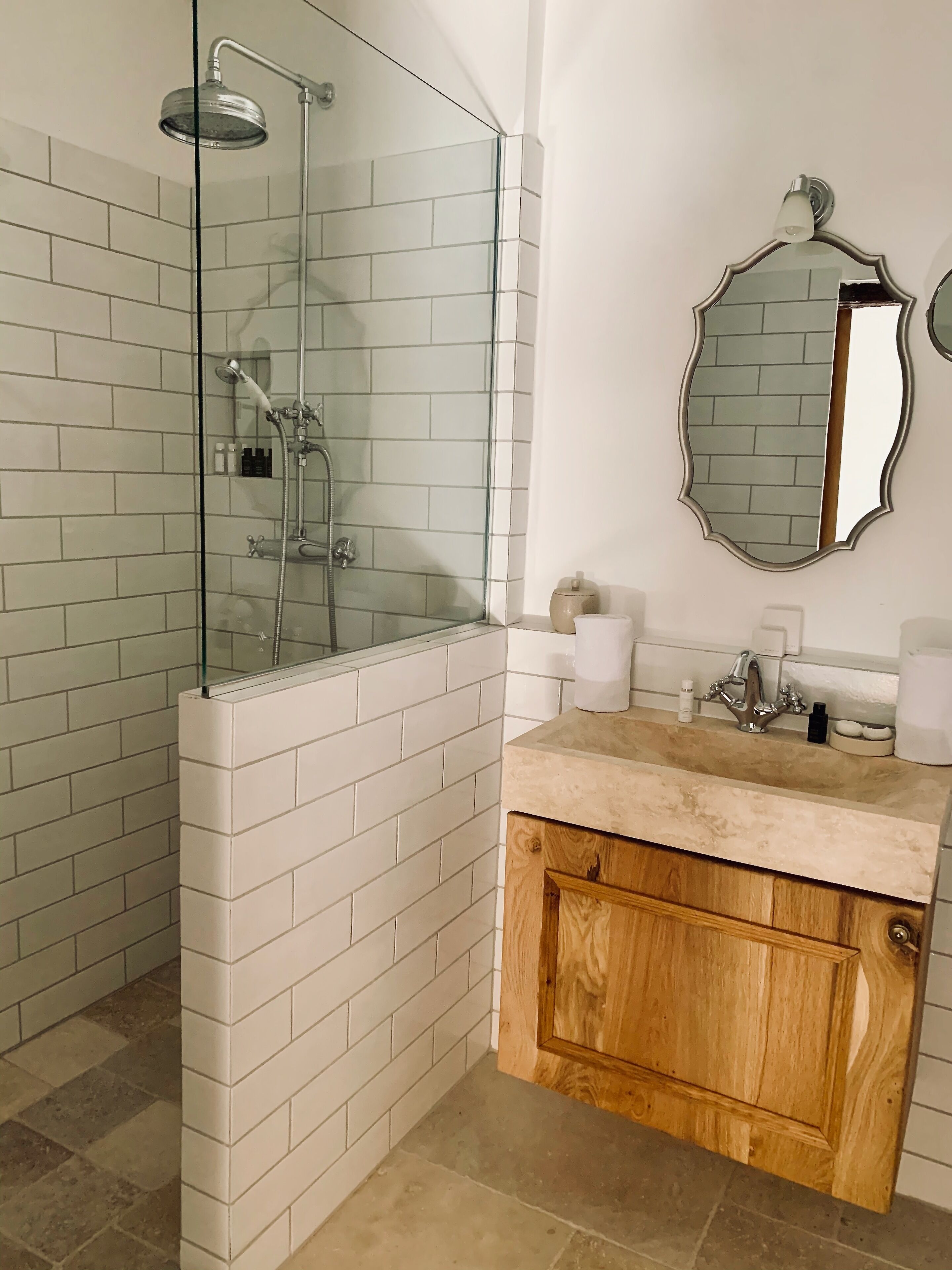 Un baño con ducha de vidrio, paredes de azulejos blancos, un tocador de madera y un espejo con un marco decorativo.
