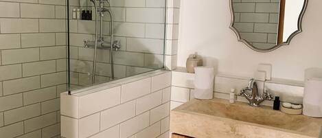 Un baño con ducha de vidrio, paredes de azulejos blancos, un tocador de madera y un espejo con un marco decorativo.