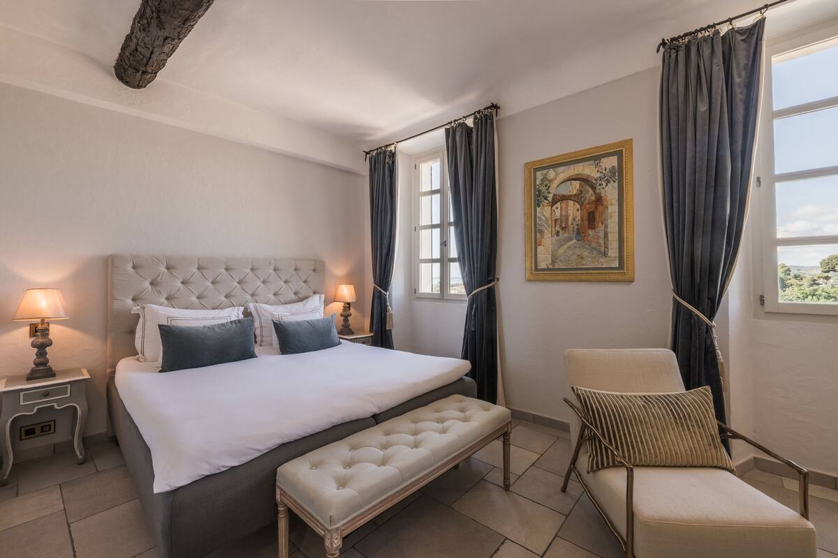 A bedroom with a large bed, a bench, a chair, and a painting on the wall.
