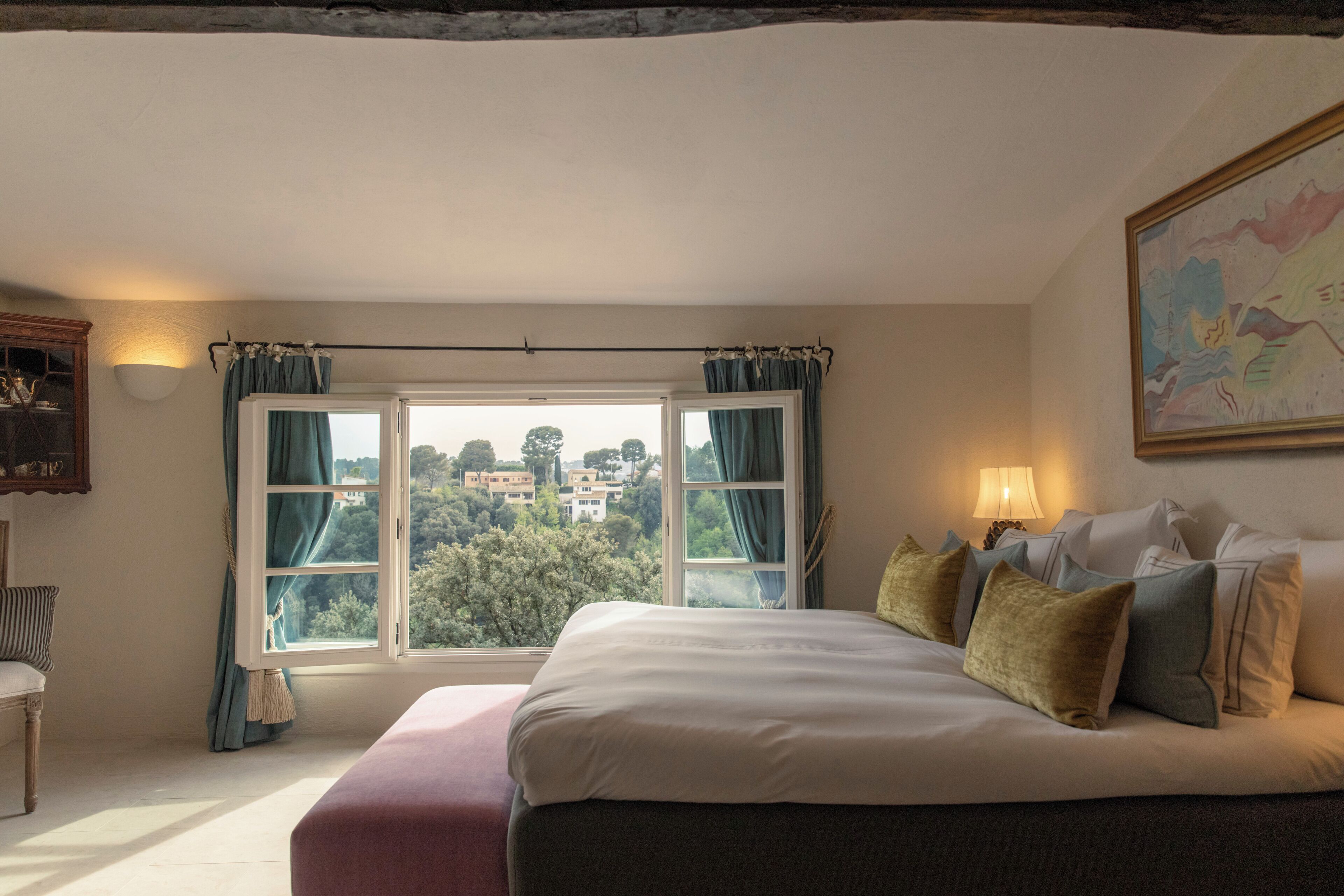 A bedroom with a large bed, a window with curtains, a lamp, and a painting on the wall.