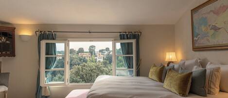 A bedroom with a large bed, a window with curtains, a lamp, and a painting on the wall.