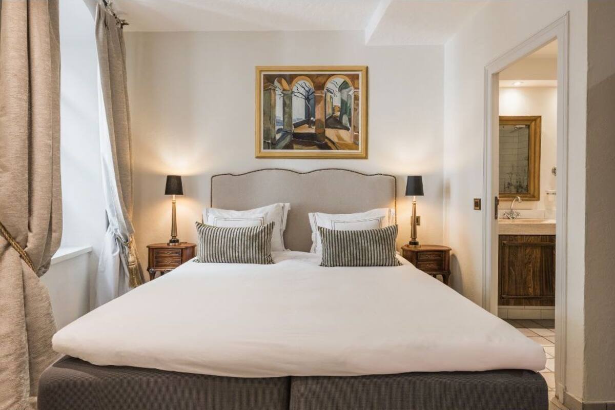 A large bed with two bedside tables, a painting above the bed, and a bathroom visible through an open door.