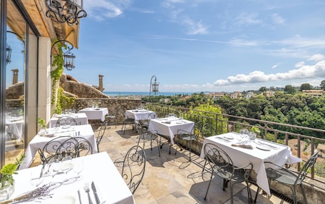 An outdoor dining area with tables and chairs, overlooking a scenic view of a coastal town and greenery.