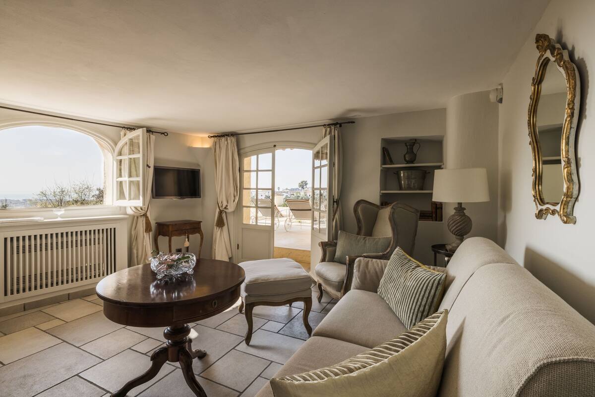 A room with a sofa, a table with a decorative item, a chair, a lamp, a mirror, and a view through a window.
