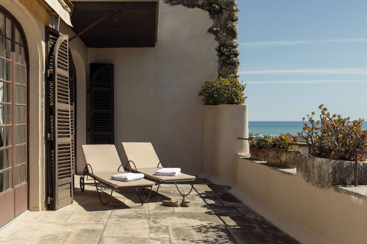 A sunlit balcony with two lounge chairs, a stone floor, and a view of the sea.