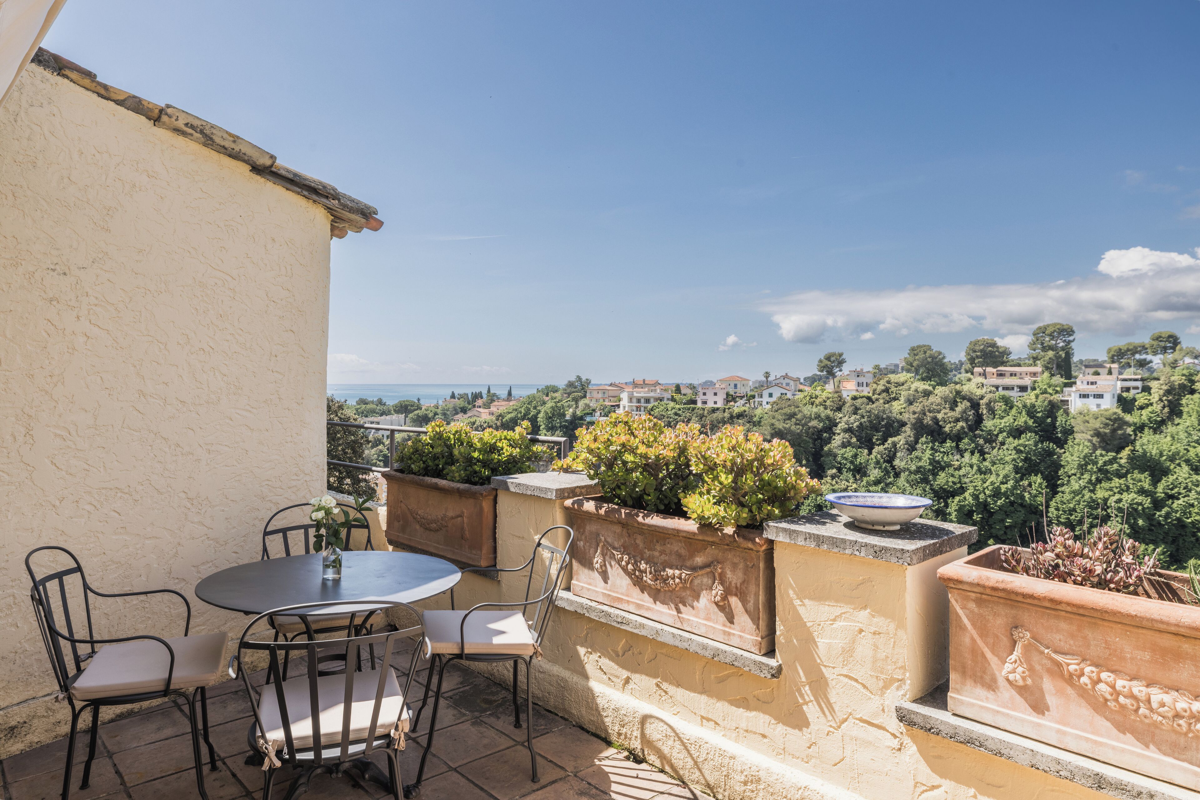 Una terraza con una mesa redonda y sillas, plantas en macetas y vistas de un paisaje urbano.