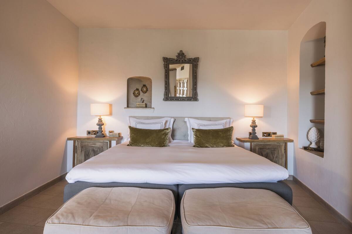 A large, neatly made bed with two pillows and two matching footstools, flanked by two bedside tables with lamps and decorative items.