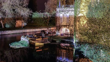 A nighttime view of a riverside park with a bridge, illuminated seating areas, and a building with arched windows.