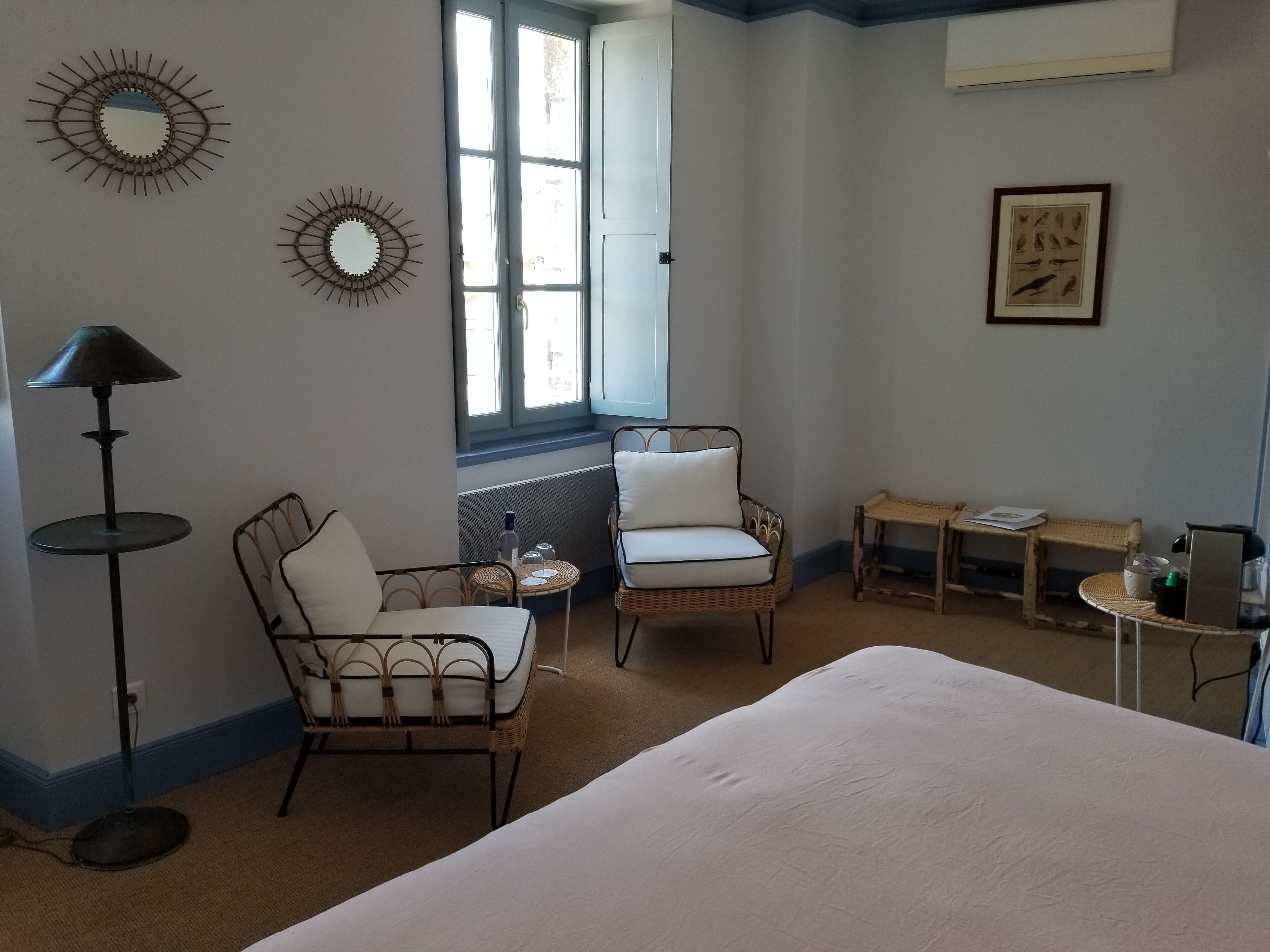Une pièce comprenant un lit, deux fauteuils en osier, une petite table, un lampadaire et des miroirs décoratifs.