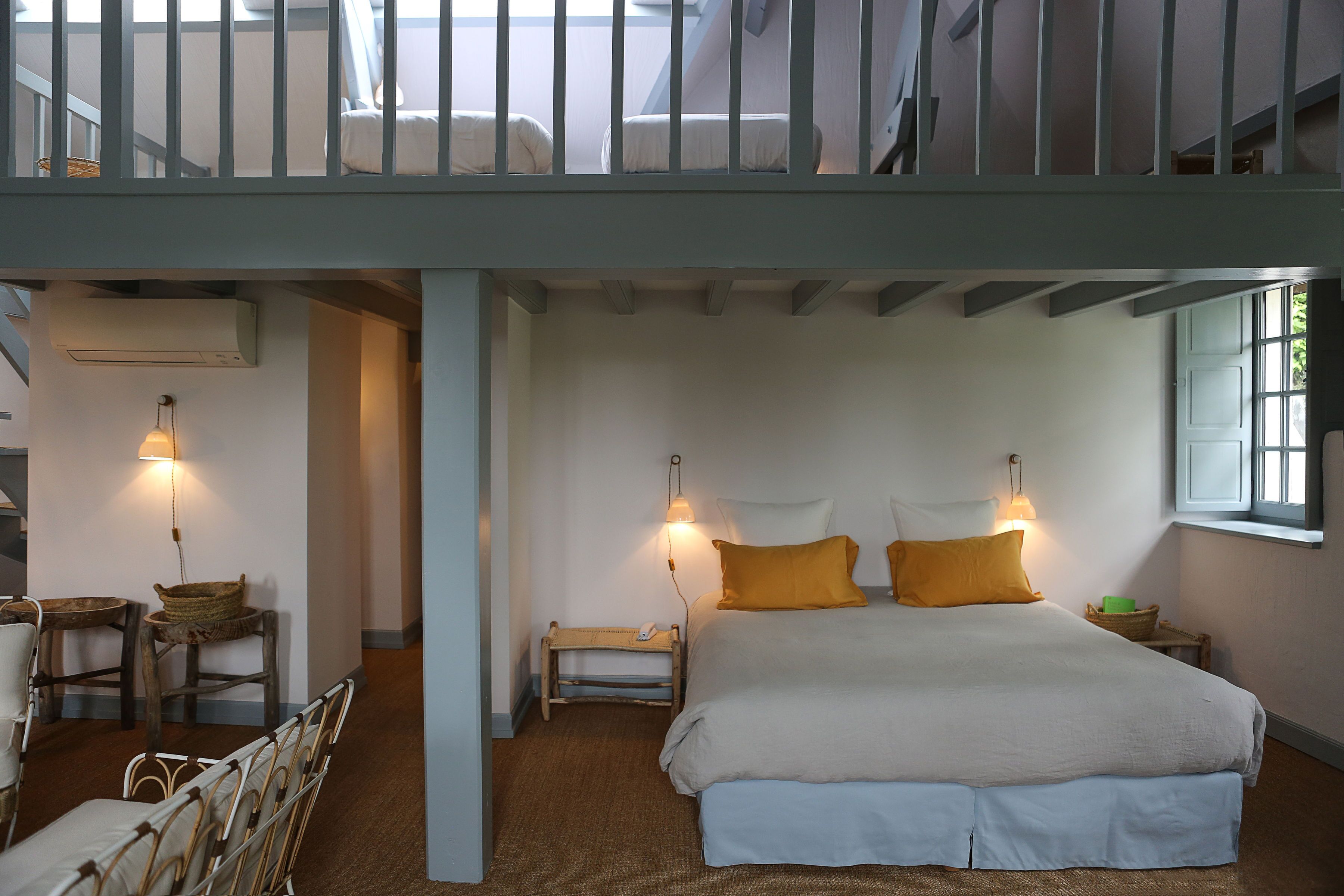 A bedroom with a bunk bed, a bedside table with a lamp, a small stool, and a window with a view of greenery.