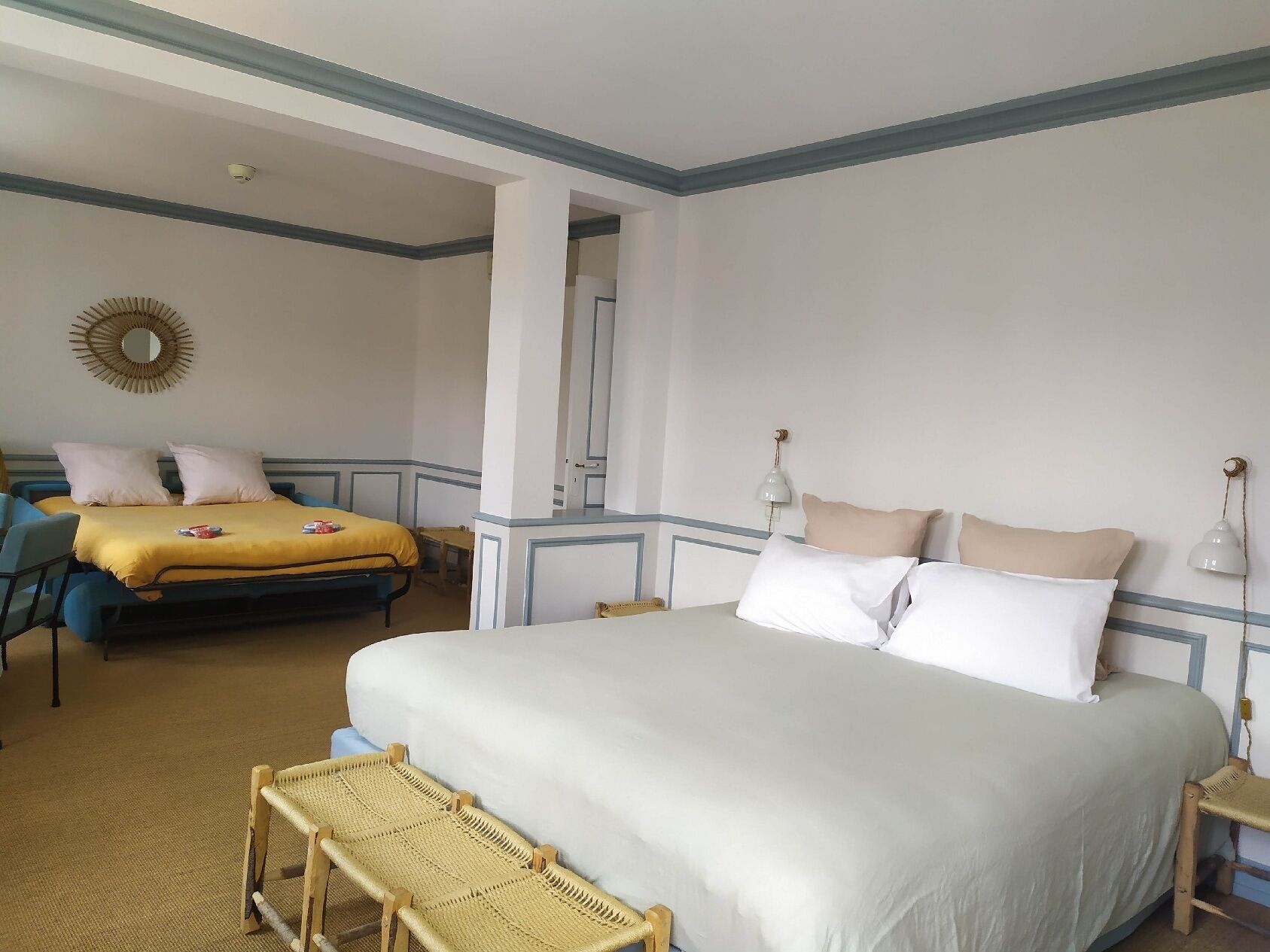 Ein Hotelzimmer mit einem großen Bett, einem kleineren Bett, zwei Stühlen, einer Bank und einem Spiegel an der Wand.
