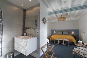 Habitación de hotel moderna con baño que incluye un tocador blanco, un espejo y un perchero. El dormitorio tiene una cama con colcha amarilla, una mesita de noche y una silla de mimbre.