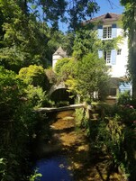 Un jardín tranquilo con un puente de piedra sobre un arroyo, abundante follaje y un edificio blanco con contraventanas azules.