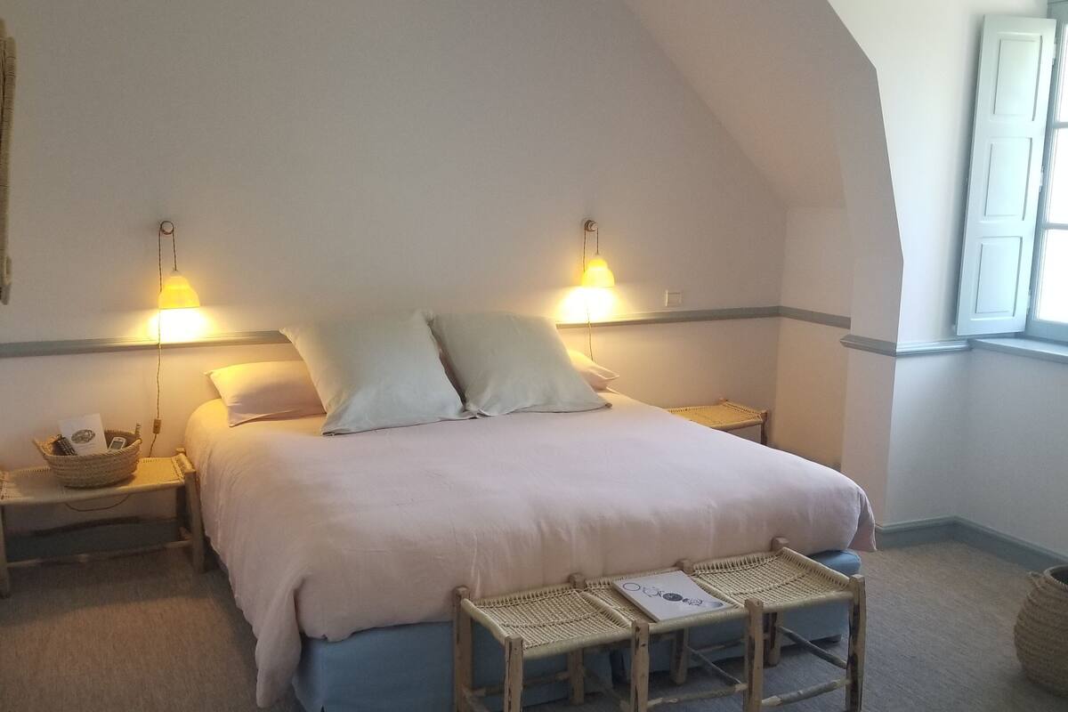 A bedroom with a large bed, two bedside lamps, a small table, and a window with curtains.