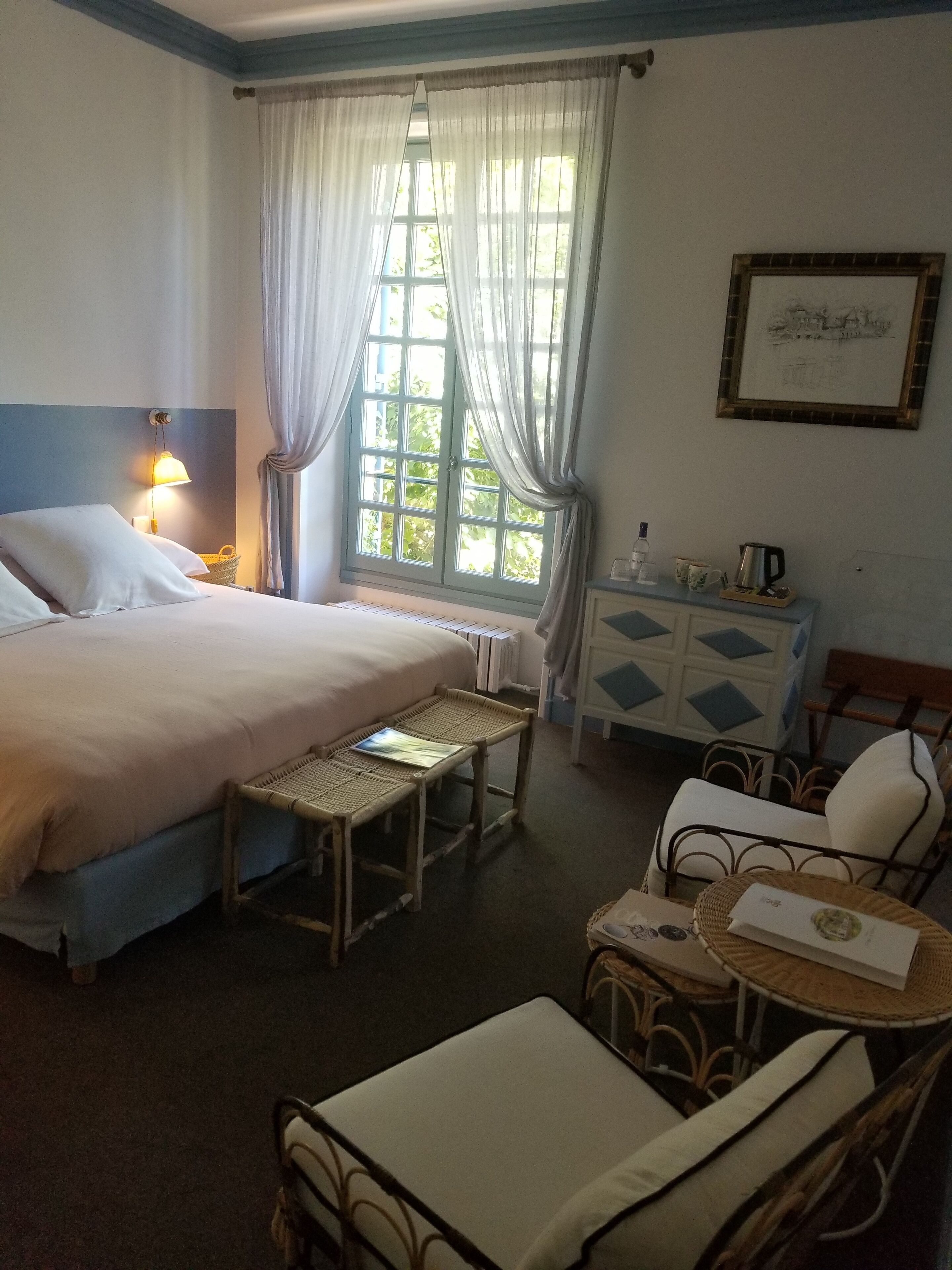 Habitación de hotel con cama, una mesita de noche con lámpara, una silla de mimbre y una ventana con cortinas.