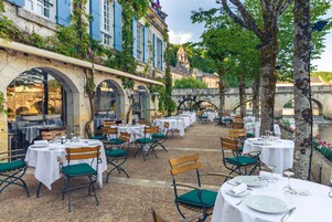 Un área de comedor al aire libre con mesas y sillas, un puente de piedra y un edificio con contraventanas azules.