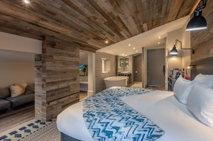 Quarto moderno com teto de madeira, uma cama grande com colcha estampada, pia embutida e um banheiro pequeno.
