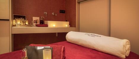 Un cuarto de spa con una toalla roja, una toalla blanca y una bandeja con velas y una botella.