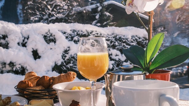 Desayuno con croissants, queso, jamón y un vaso de jugo de naranja, con montañas al fondo.
