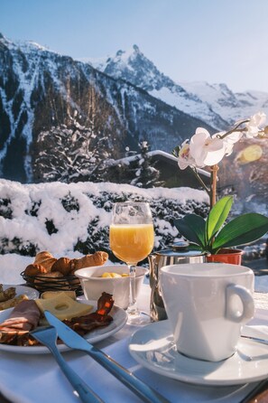 Desayuno con croissants, queso, jamón y un vaso de jugo de naranja, con montañas al fondo.