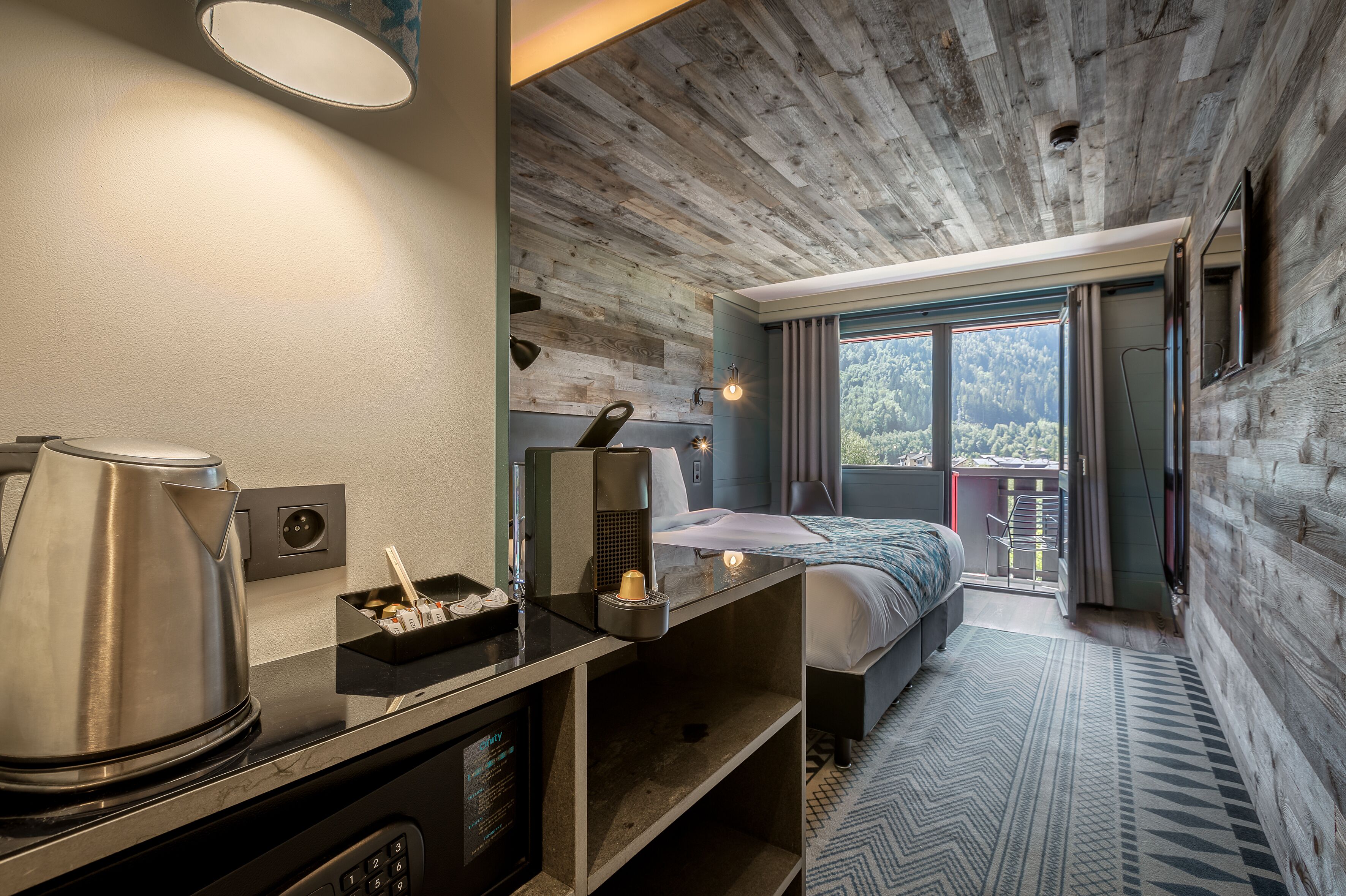 Een moderne hotelkamer met een houten plafond, een bed, een bureau met waterkoker en uitzicht op het omliggende landschap.