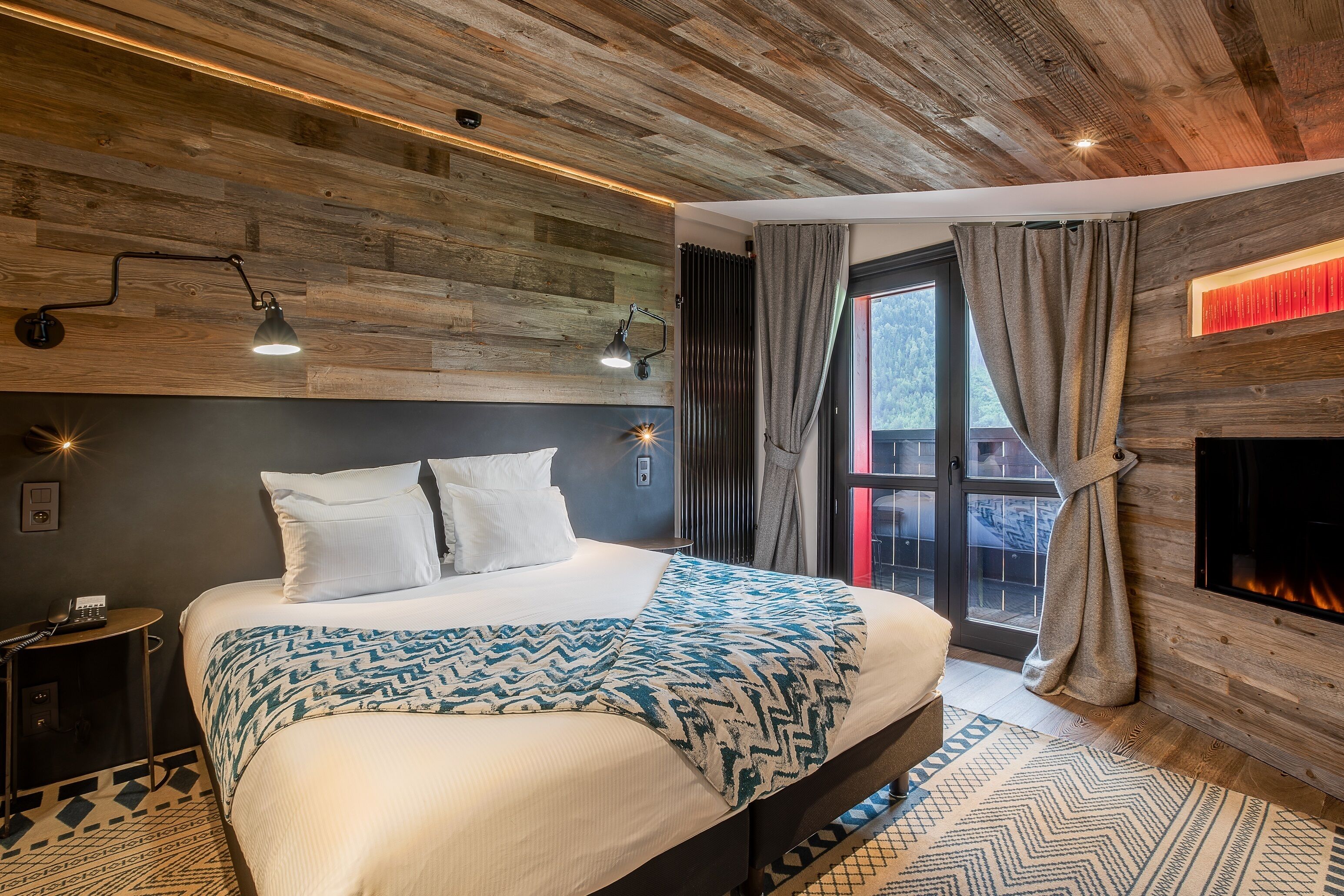 Un dormitorio moderno con techo de madera, una cama grande con ropa de cama estampada, una mesita de noche con lámpara y una chimenea.