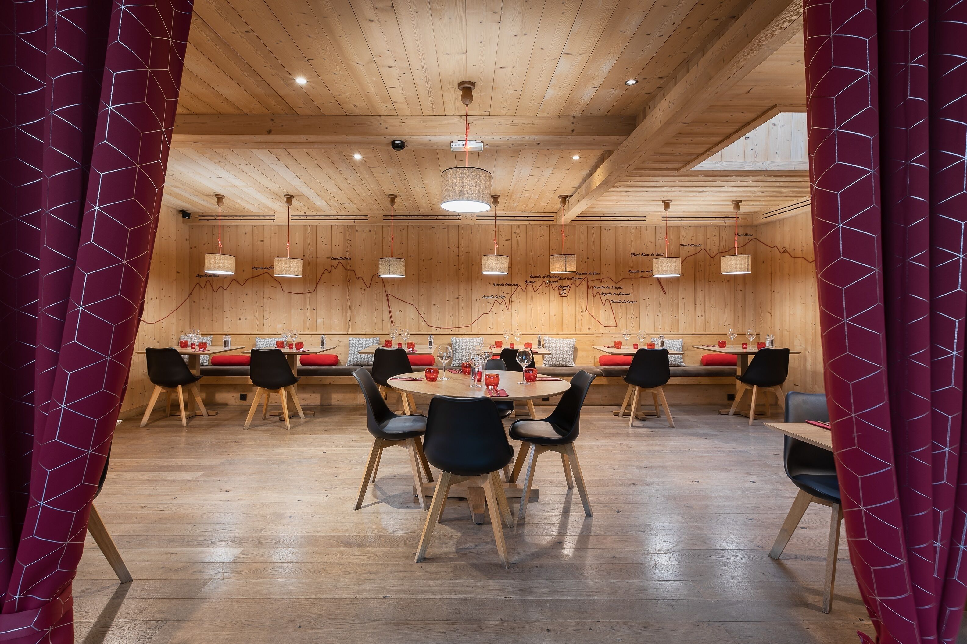 A wooden interior with round tables and black chairs, featuring a ceiling with wooden beams and pendant lights.