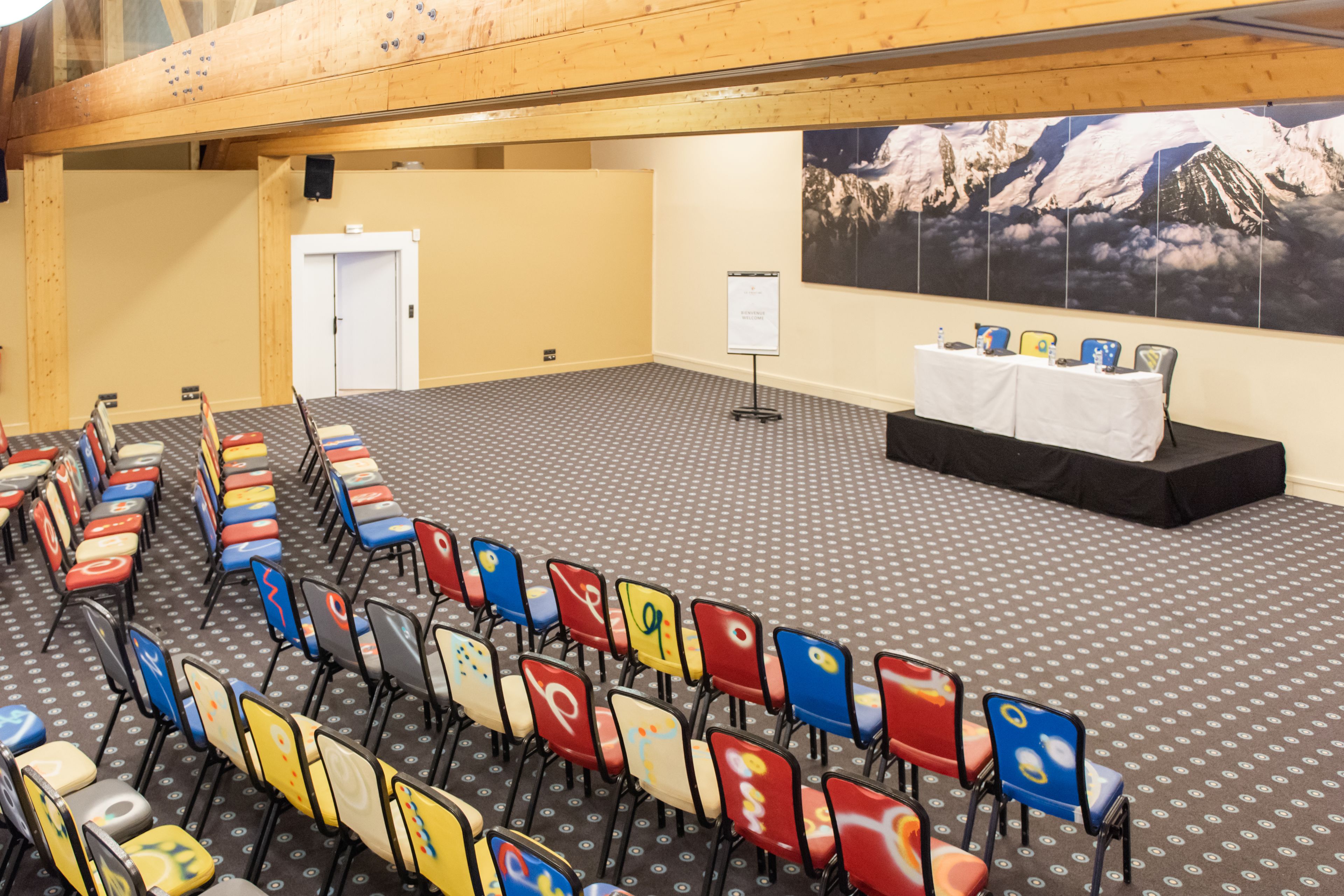 Une salle de conférence avec des rangées de chaises, une scène surmontée d’une table blanche et une grande affiche représentant un paysage de montagne accrochée au mur.