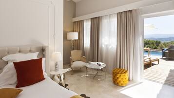 Een hotelkamer met een bed, een stoel, een klein tafeltje, een lamp en uitzicht op een zwembad en bergen.